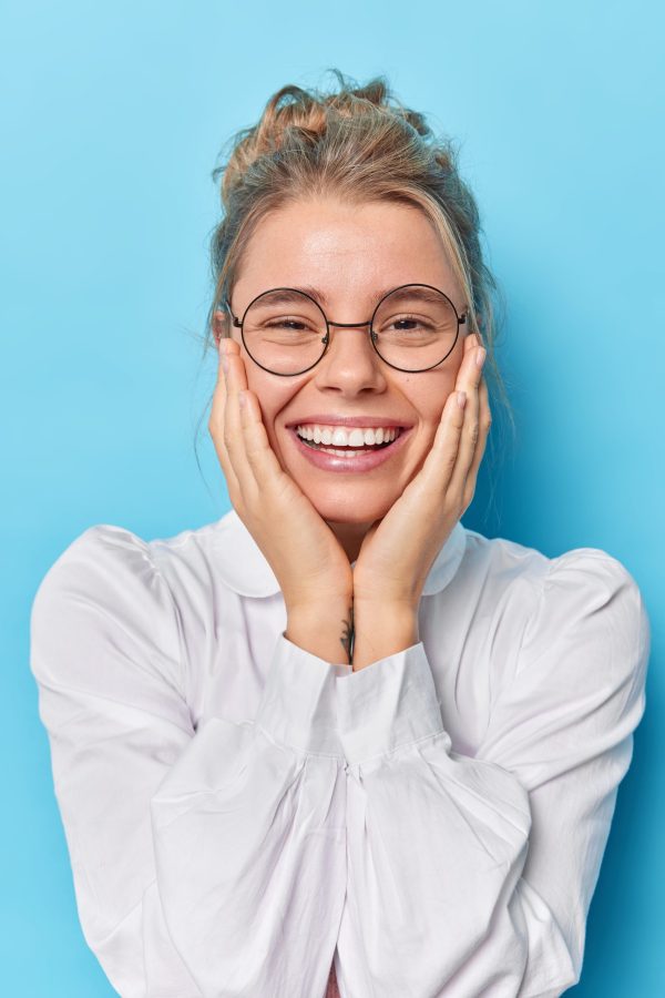 Good looking lovely European woman keeps both hands on cheeks smiles broadly dressed in white shirt transparent eyeglasses hears pleasant news poses against blue background looks cute at camera