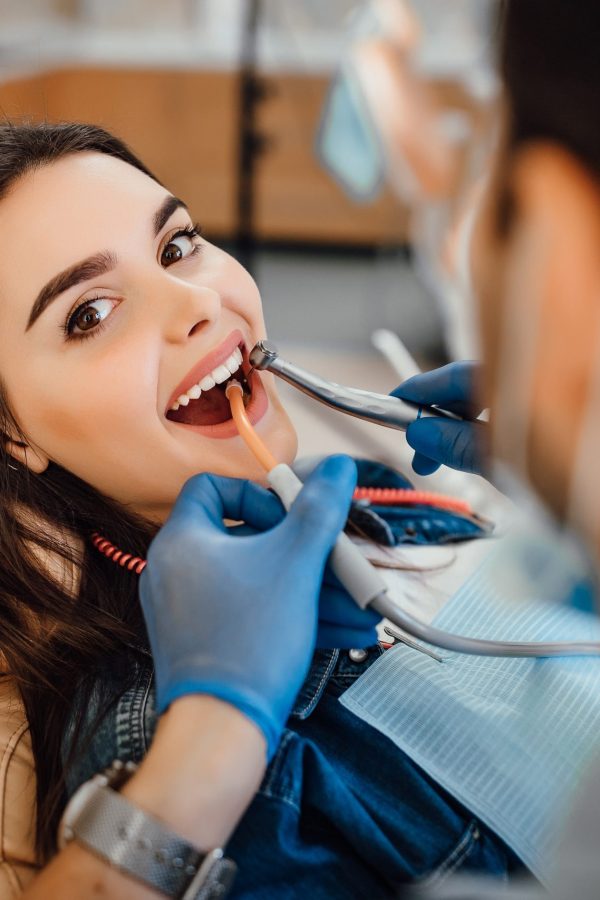 Young female patient visiting dentist office. Beautiful woman with healthy straight white teeth sitting at dental chair with open mouth during oral checkup while doctor working at teeth.